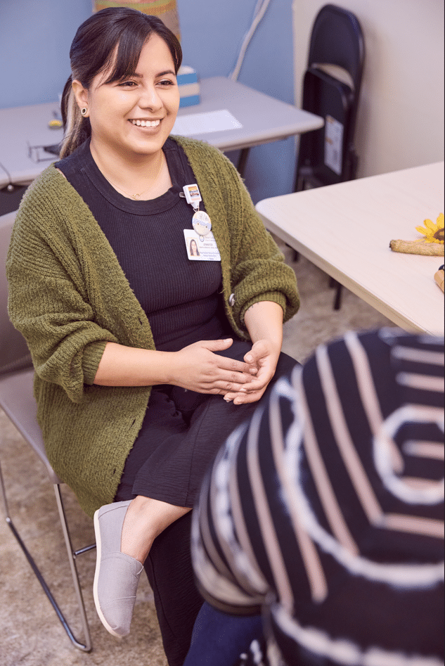 A Smiling Woman In An Olive Green Cardigan And A Name Tag Sits In A Chair, Talking To Someone Whose Back Is Turned To The Camera.